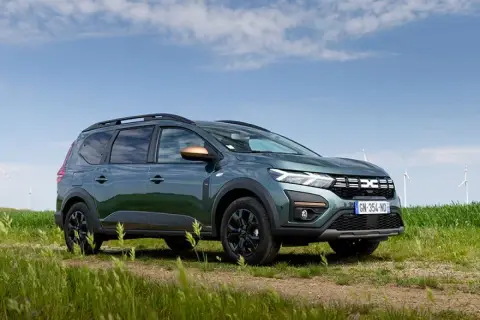 Un'auto familiare verde scuro, con barre sul tetto e dettagli color rame, parcheggiata su un sentiero sterrato in un campo con mulini a vento sullo sfondo.