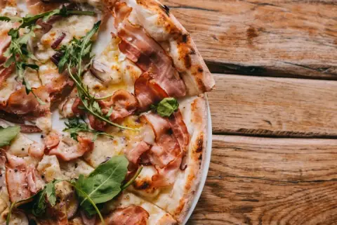 Pyszna pizza carbonara z chrupiącym boczkiem i rukolą, podana na drewnianym stole.