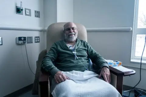 Uśmiechnięty mężczyzna w fotelu szpitalnym, podłączony do kroplówki, otrzymuje żywienie pozajelitowe.