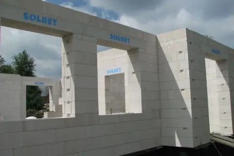 Budowa domu z bloczków SOLBET. Widoczny mur z otworami okiennymi i drzwiami, w tle drzewa i niebo.