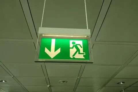 Zielone oświetlenie ewakuacyjne z ikoną strzałki w dół i biegnącą postacią, wiszące pod sufitem.