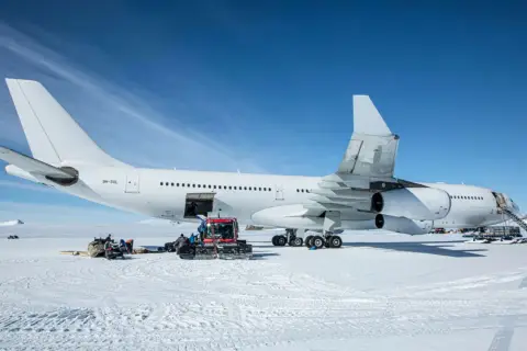 Biały airbus a340-300 na śniegu, z obsługą naziemną i ratrakiem.