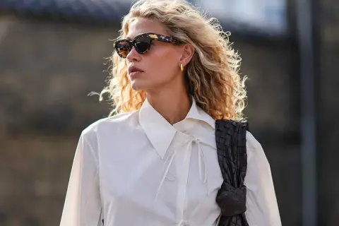 Eine skandinavische Blonde mit lockigem Haar, Sonnenbrille und weißem Hemd, die eine dunkle Tasche trägt.