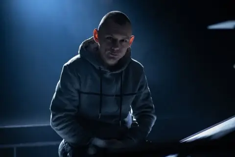 Młody polski raper w szarej bluzie z kapturem, oświetlony od tyłu niebieskim światłem.