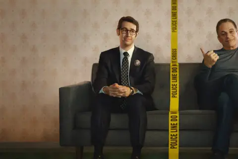 Dwóch mężczyzn siedzi na kanapie. Jeden, ubrany w garnitur, to **the good cop**. Obok niego, za taśmą policyjną, drugi mężczyzna uśmiecha się.