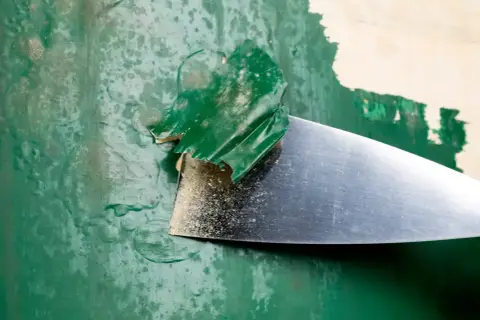 Metalowa szpachelka zdziera starą, zieloną farbę, odsłaniając podkład.