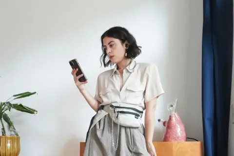 Kobieta w beżowej koszuli i szarej spódnicy z saszetką na biodrach przegląda telefon, jakby szukała okazji na Vinted pro.