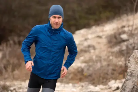 Mężczyzna w niebieskiej kurtce softshell i czapce biegnie po kamienistym terenie. Pamiętaj, jak prać softshell, by służył Ci latami.