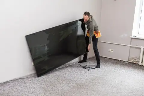 Mężczyzna z pasem narzędziowym przenosi duży, czarny ekran. To telewizor o wymiarach 55 cali, gotowy do montażu.