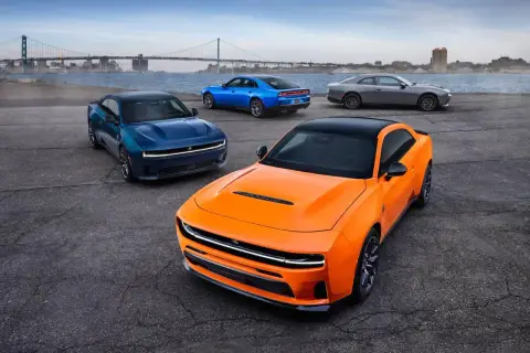 Pomarańczowy i niebieski Dodge Charger Hellcat oraz szary coupe stoją nad wodą z mostem w tle.
