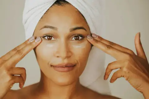 Mujer con toalla en la cabeza aplica crema con ceramida alrededor de sus ojos.
