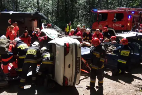 Akcja ratunkowa ksrg po wypadku. Strażacy wyciągają poszkodowanych z przewróconego samochodu. W tle wozy strażackie i karetka.