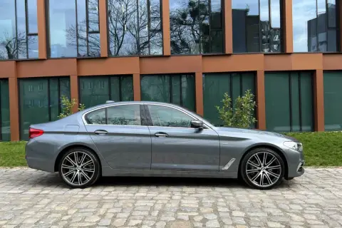 Szary sedan BMW M5 G30 stoi na brukowanym podjeździe przed nowoczesnym budynkiem z brązowymi ramami okien.