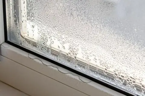 Gotas de agua cubren el cristal de la ventana, indicando condensación. El alféizar muestra pequeñas acumulaciones de agua.