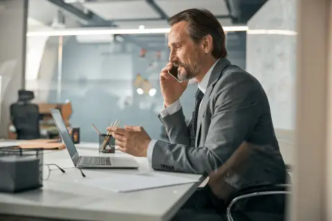 Mężczyzna w garniturze rozmawia przez telefon, patrząc na laptopa. To typowy dzień pracy CEO, co to znaczy? To znaczy, że zarządza firmą.
