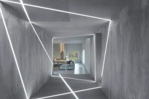 Nowoczesny korytarz z betonowymi ścianami i geometrycznym podświetleniem ściany od góry. W tle salon z kominkiem i kanapą.