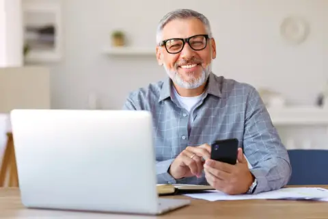 Uśmiechnięty senior z okularami pracuje przy laptopie i telefonie. To ulga dla pracujących seniorów, którzy cenią sobie nowoczesne rozwiązania.