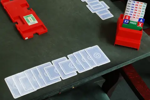 Stół do gry w brydża. Rozłożone karty, czerwone pudełko z oznaczeniami licytacji i kartami "PASS", "XX", "A". Gra karciana dla dwóch par z licytacją.