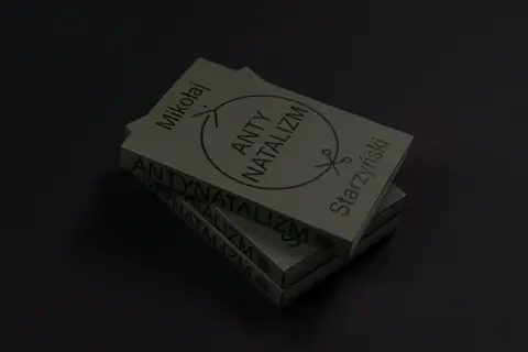 Stos książek z napisem "ANTY NATALIZM" i symbolem nożyczek. Dzieło Mikołaja Starzyńskiego.