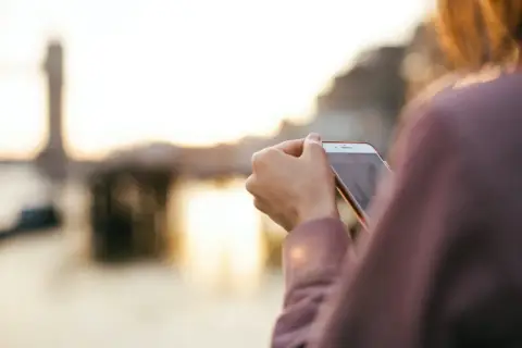 Osoba sprawdza na telefonie, jak włączyć roaming w Orange na kartę, podczas zachodu słońca nad wodą.