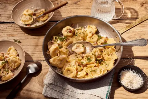 Herzhafte Tortellini Carbonara, garniert mit Petersilie und Parmesan, serviert in einer Pfanne und Schalen auf Holztisch.