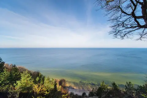 Widok z **wzgórza Gosań** na spokojne morze i piaszczystą plażę. Na pierwszym planie drzewa i zieleń.