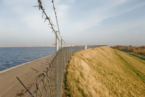 Zapora wodna przy elektrowni w Żarnowcu, z drutem kolczastym i ogrodzeniem. Po lewej woda, po prawej trawiasty nasyp.