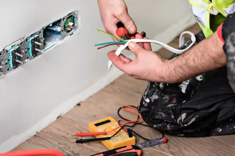 Elektryk przygotowuje kabel czarny, brązowy, niebieski do podłączenia gniazdek. Narzędzia i miernik leżą na podłodze.