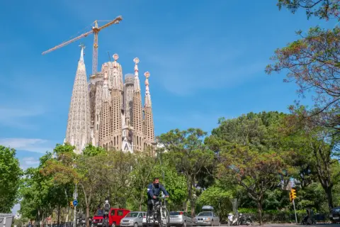 Majestatyczna La Sagrada Familia w Barcelonie, wciąż w budowie, góruje nad zielonym parkiem i ulicą, gdzie rowerzysta przemierza drogę.