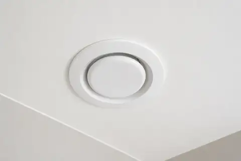 Biały, okrągły anemostat wywiewny zamontowany na białym suficie.