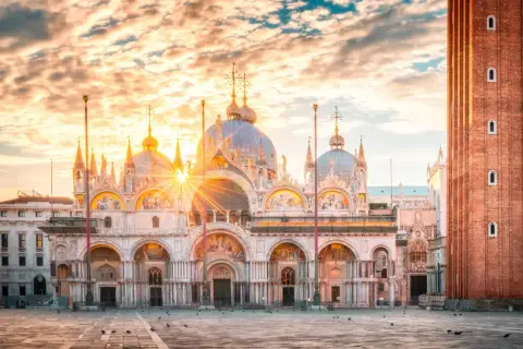 Wschód słońca nad Bazyliką św. Marka na Piazza San Marco. Złote promienie przebijają się przez chmury, oświetlając wenecką architekturę.