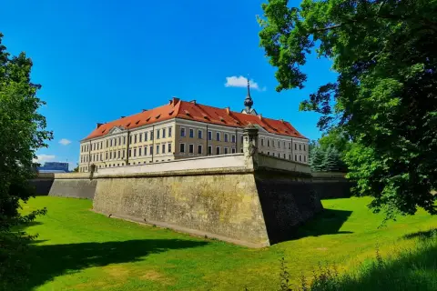 Zamek w Rzeszowie z czerwonym dachem i fosą otoczoną zielonym trawnikiem, pod błękitnym niebem.