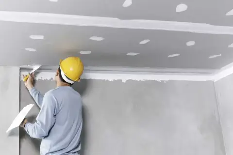 Pracownik w żółtym kasku nakłada gładź na sufit. To etap prac wykończeniowych, gdzie przygotowywany jest styropian na sufit w pokoju.