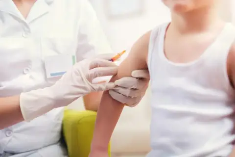 Dziecko otrzymuje szczepionkę 6w1. Pielęgniarka w białym fartuchu i rękawiczkach podaje zastrzyk w ramię malucha.