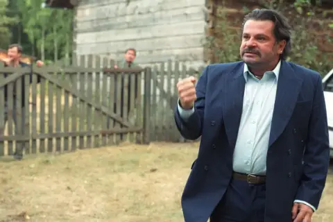 Mężczyzna w garniturze, jakby z filmu "To ja, złodziej", pokazuje pięść. Za nim przez płot wyglądają inni.