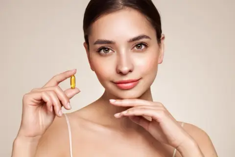 Mujer sonriente sostiene una cápsula dorada de vitamina e, símbolo de cuidado y bienestar para una piel radiante.