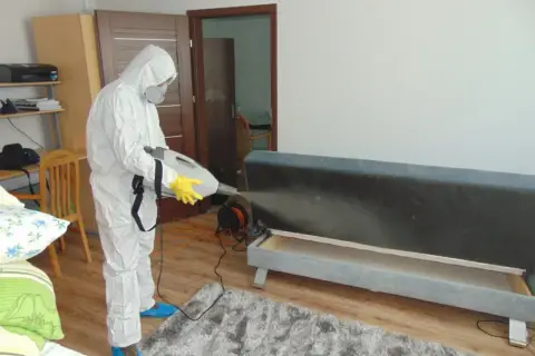 Osoba w kombinezonie ochronnym wykonuje dezynsekcję, co to jest? Zabieg z użyciem opryskiwacza na kanapie.