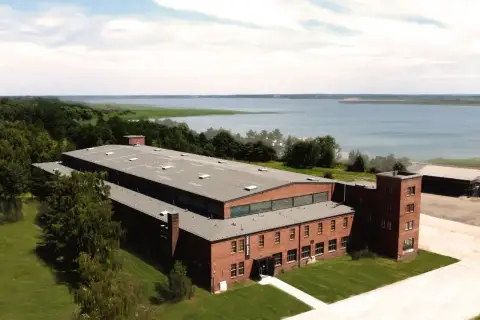 Dawny kompleks przemysłowy, dziś arche Mielno, z ceglanymi budynkami i wieżą, nad jeziorem.