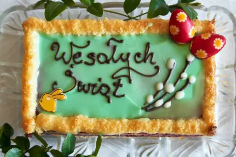 Zielone ciasto wielkanocne z napisem "Wesołych Świąt", ozdobione zajączkiem, baziami i pisankami.