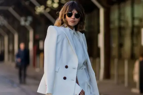 Mujer con traje blanco, gafas de sol redondas y cabello castaño. Su atuendo, elegante y sencillo, refleja el **minimalista significado** de la moda.