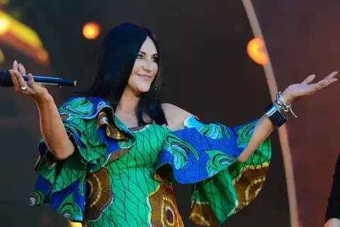 Kayah w kolorowej sukience z falbanami śpiewa na scenie, trzymając mikrofon.