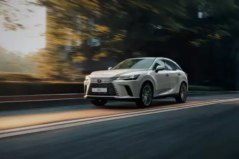 Biały Lexus RX 350h pędzi po drodze, otoczony rozmazanymi drzewami i światłem zachodzącego słońca.