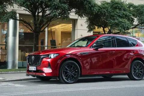 Lśniąca, czerwona Mazda CX-60 3.3 diesel na tle nowoczesnego budynku i drzew.