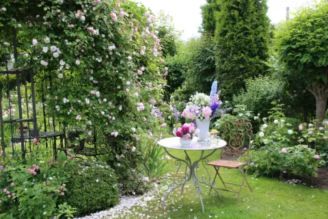 Ein romantischer Garten mit blühenden Rosen, ein Tisch mit Blumenarrangements und ein schmiedeeiserner Stuhl laden zum Verweilen ein. Ein idyllischer Ort, um einen **beet anlegen**-Traum zu verwirklichen.