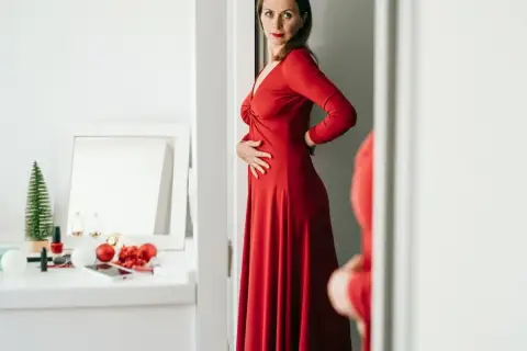 Kobieta w czerwonej, eleganckiej sukience, która optycznie wyszczupla. Na toaletce świąteczne ozdoby i kosmetyki.