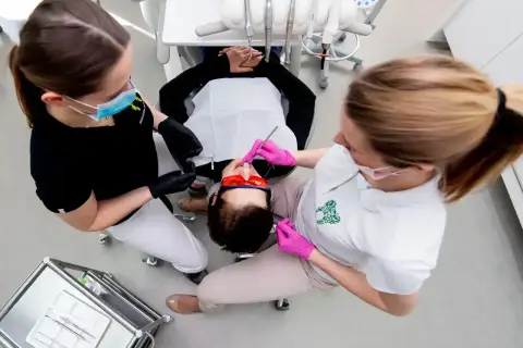 Dwie asystentki stomatologiczne pracują przy pacjencie. Jedna z nich, w czarnej bluzce, przygotowuje narzędzia. Druga, w białej koszulce, wykonuje zabieg.