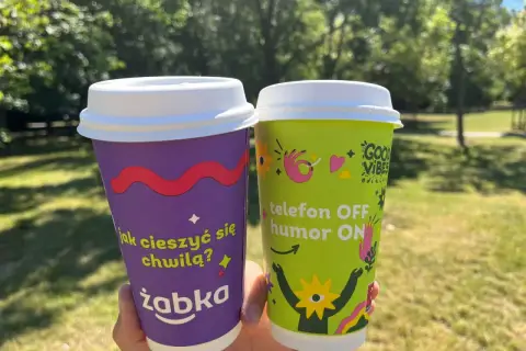 Dwie kawy na wynos z Żabki. Fioletowy kubek pyta "jak cieszyć się chwilą?", zielony głosi "telefon OFF, humor ON".