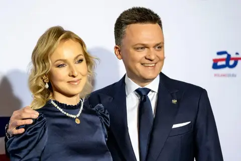 Szymon Hołownia z żoną, uśmiechnięci, pozują do zdjęcia. Ona w granatowej sukience i perłach, on w garniturze.