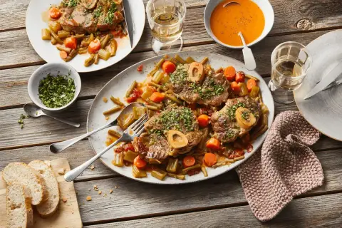 Ein festliches Mahl: saftiges Ossobuco mit Gemüse und einem Glas Weißwein. Dazu frisches Brot und eine cremige Tomatensuppe.