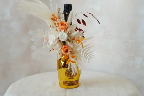 Goldene Weinflasche mit Blumen verpackt. Ein Strauß aus getrockneten Blumen, Rosen und Pampasgras schmückt die Flasche.